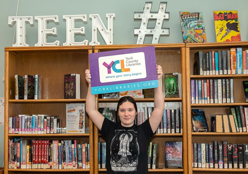 teen holding york county libraries sign in front of bookshelf with a "hashtag teen" sign on top of the shelf