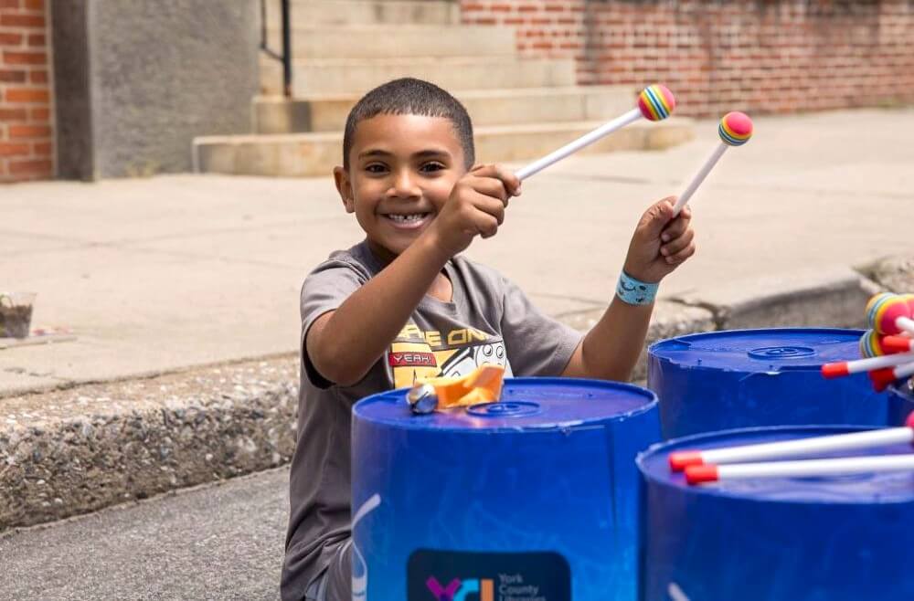 little boy smiling at the camera while playing a bucket drum.