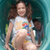 little girl sitting at the edge of a playground crawl tube wearing the summerquest 2025 color our world t-shirt. shirt has a colorful illustration of kids with a rainbow behind them and the text "color our world" to the left.