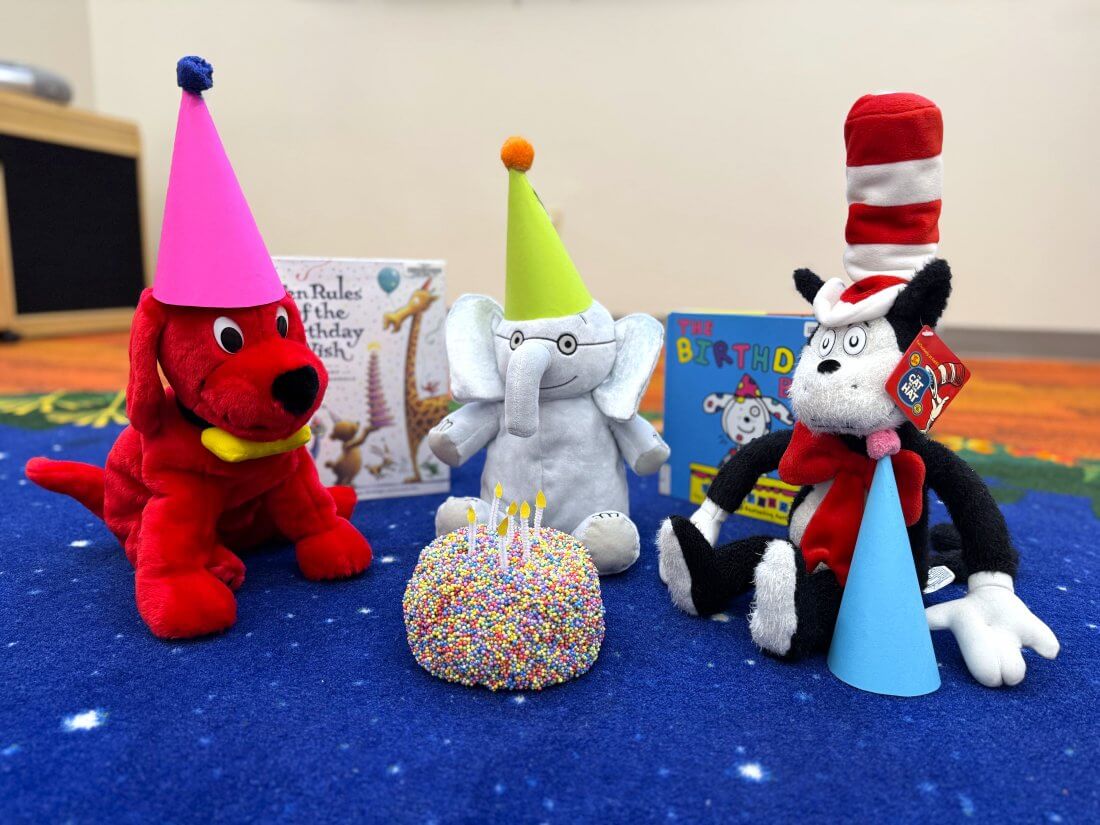 stuffed animals wearing birthday party hats while sitting on the ground around a toy birthday cake