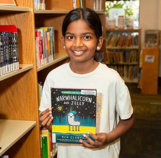 little girl holding a book up smiling at camera